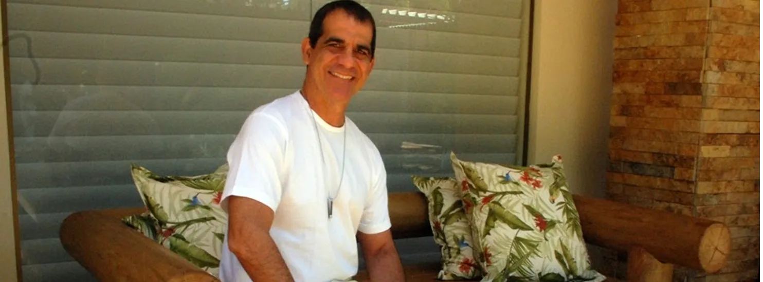 Durval Lelys junto a banco de madeira roliça e parede revestida de pedra em sua residência em Busca Vida
