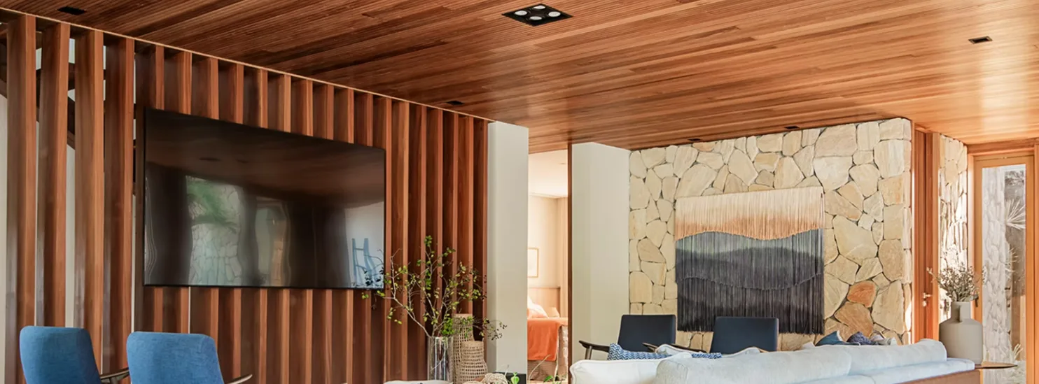 Living com forro e painel ripado de madeira junto à parede de pedra na Casa Praia Bella